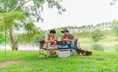 İki genç Asyalı erkek, kamp alanında dinlenirken tablet üzerinde video oyunu oynamaktan hoşlanırlar. Kamp malzemeleri, gitarlar ve doğa ile çevrili sahne, modern ve neşeli doğa deneyimini yakalar..