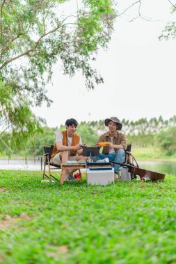 İki genç Asyalı erkek, kamp alanında dinlenirken tablet üzerinde video oyunu oynamaktan hoşlanırlar. Kamp malzemeleri, gitarlar ve doğa ile çevrili sahne, modern ve neşeli doğa deneyimini yakalar..