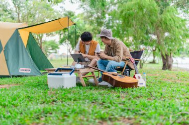 İki genç Asyalı erkek, kamp alanında dinlenirken tablet üzerinde video oyunu oynamaktan hoşlanırlar. Kamp malzemeleri, gitarlar ve doğa ile çevrili sahne, modern ve neşeli doğa deneyimini yakalar..
