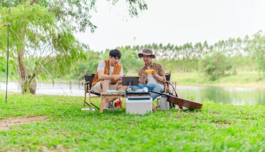 İki genç Asyalı erkek, kamp alanında dinlenirken tablet üzerinde video oyunu oynamaktan hoşlanırlar. Kamp malzemeleri, gitarlar ve doğa ile çevrili sahne, modern ve neşeli doğa deneyimini yakalar..