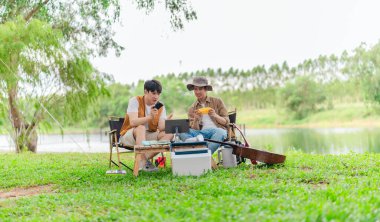 İki genç Asyalı erkek, kamp alanında dinlenirken tablet üzerinde video oyunu oynamaktan hoşlanırlar. Kamp malzemeleri, gitarlar ve doğa ile çevrili sahne, modern ve neşeli doğa deneyimini yakalar..