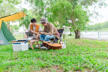 İki genç Asyalı erkek, kamp alanında dinlenirken tablet üzerinde video oyunu oynamaktan hoşlanırlar. Kamp malzemeleri, gitarlar ve doğa ile çevrili sahne, modern ve neşeli doğa deneyimini yakalar..