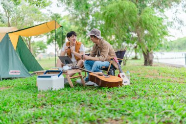 İki genç Asyalı erkek, kamp alanında dinlenirken tablet üzerinde video oyunu oynamaktan hoşlanırlar. Kamp malzemeleri, gitarlar ve doğa ile çevrili sahne, modern ve neşeli doğa deneyimini yakalar..