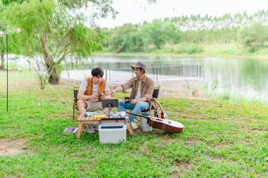 İki genç Asyalı erkek, kamp alanında dinlenirken tablet üzerinde video oyunu oynamaktan hoşlanırlar. Kamp malzemeleri, gitarlar ve doğa ile çevrili sahne, modern ve neşeli doğa deneyimini yakalar..