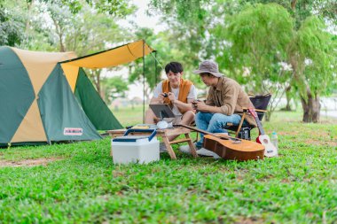 İki genç Asyalı erkek, kamp alanında dinlenirken tablet üzerinde video oyunu oynamaktan hoşlanırlar. Kamp malzemeleri, gitarlar ve doğa ile çevrili sahne, modern ve neşeli doğa deneyimini yakalar..