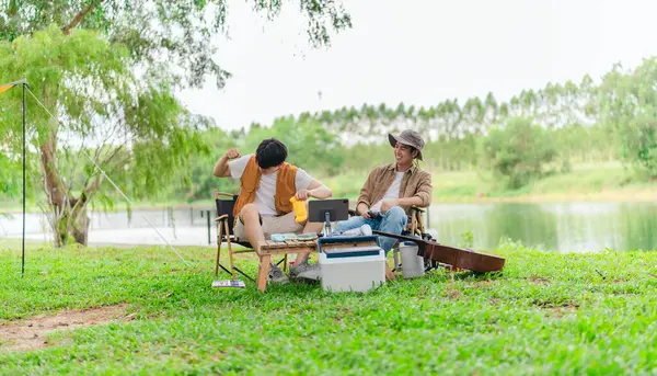 İki genç Asyalı erkek, kamp alanında dinlenirken tablet üzerinde video oyunu oynamaktan hoşlanırlar. Kamp malzemeleri, gitarlar ve doğa ile çevrili sahne, modern ve neşeli doğa deneyimini yakalar..