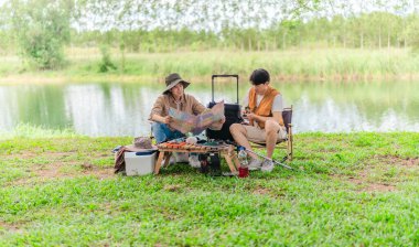 İki Asyalı erkek arkadaş, doğa ve teçhizatla çevrili bir gölün kenarında sakin bir kamp deneyimi yaşıyor. Sahne, rahatlamış bir doğa keşfi anı yakalıyor ve manzaralı bir ortamda hazırlanıyor..