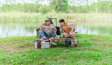 İki Asyalı erkek arkadaş, doğa ve teçhizatla çevrili bir gölün kenarında sakin bir kamp deneyimi yaşıyor. Sahne, rahatlamış bir doğa keşfi anı yakalıyor ve manzaralı bir ortamda hazırlanıyor..