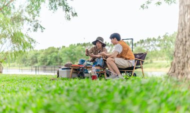 İki Asyalı erkek arkadaş, doğa ve teçhizatla çevrili bir gölün kenarında sakin bir kamp deneyimi yaşıyor. Sahne, rahatlamış bir doğa keşfi anı yakalıyor ve manzaralı bir ortamda hazırlanıyor..