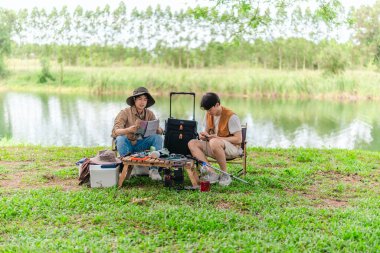 İki Asyalı erkek arkadaş, doğa ve teçhizatla çevrili bir gölün kenarında sakin bir kamp deneyimi yaşıyor. Sahne, rahatlamış bir doğa keşfi anı yakalıyor ve manzaralı bir ortamda hazırlanıyor..