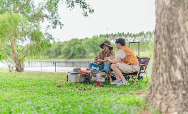 İki Asyalı erkek arkadaş, doğa ve teçhizatla çevrili bir gölün kenarında sakin bir kamp deneyimi yaşıyor. Sahne, rahatlamış bir doğa keşfi anı yakalıyor ve manzaralı bir ortamda hazırlanıyor..