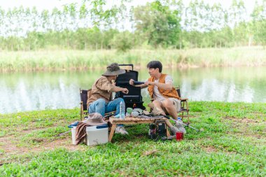 İki Asyalı erkek arkadaş, doğa ve teçhizatla çevrili bir gölün kenarında sakin bir kamp deneyimi yaşıyor. Sahne, rahatlamış bir doğa keşfi anı yakalıyor ve manzaralı bir ortamda hazırlanıyor..