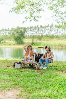 İki Asyalı erkek arkadaş, doğa ve teçhizatla çevrili bir gölün kenarında sakin bir kamp deneyimi yaşıyor. Sahne, rahatlamış bir doğa keşfi anı yakalıyor ve manzaralı bir ortamda hazırlanıyor..