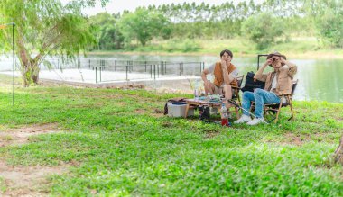 İki Asyalı erkek arkadaş, doğa ve teçhizatla çevrili bir gölün kenarında sakin bir kamp deneyimi yaşıyor. Sahne, rahatlamış bir doğa keşfi anı yakalıyor ve manzaralı bir ortamda hazırlanıyor..