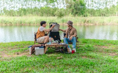 İki Asyalı erkek arkadaş, doğa ve teçhizatla çevrili bir gölün kenarında sakin bir kamp deneyimi yaşıyor. Sahne, rahatlamış bir doğa keşfi anı yakalıyor ve manzaralı bir ortamda hazırlanıyor..