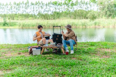 İki Asyalı erkek arkadaş, doğa ve teçhizatla çevrili bir gölün kenarında sakin bir kamp deneyimi yaşıyor. Sahne, rahatlamış bir doğa keşfi anı yakalıyor ve manzaralı bir ortamda hazırlanıyor..