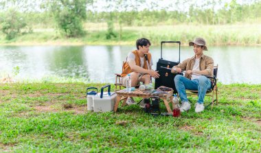 İki Asyalı erkek arkadaş, doğa ve teçhizatla çevrili bir gölün kenarında sakin bir kamp deneyimi yaşıyor. Sahne, rahatlamış bir doğa keşfi anı yakalıyor ve manzaralı bir ortamda hazırlanıyor..