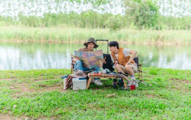 İki Asyalı erkek arkadaş, doğa ve teçhizatla çevrili bir gölün kenarında sakin bir kamp deneyimi yaşıyor. Sahne, rahatlamış bir doğa keşfi anı yakalıyor ve manzaralı bir ortamda hazırlanıyor..