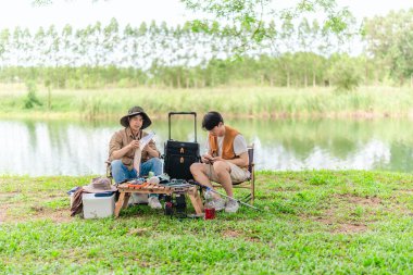 İki Asyalı erkek arkadaş, doğa ve teçhizatla çevrili bir gölün kenarında sakin bir kamp deneyimi yaşıyor. Sahne, rahatlamış bir doğa keşfi anı yakalıyor ve manzaralı bir ortamda hazırlanıyor..