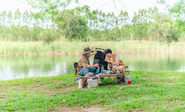İki Asyalı erkek arkadaş, doğa ve teçhizatla çevrili bir gölün kenarında sakin bir kamp deneyimi yaşıyor. Sahne, rahatlamış bir doğa keşfi anı yakalıyor ve manzaralı bir ortamda hazırlanıyor..