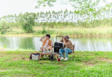 İki Asyalı erkek arkadaş, doğa ve teçhizatla çevrili bir gölün kenarında sakin bir kamp deneyimi yaşıyor. Sahne, rahatlamış bir doğa keşfi anı yakalıyor ve manzaralı bir ortamda hazırlanıyor..