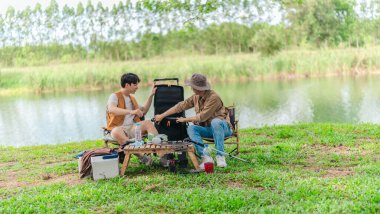 İki Asyalı erkek arkadaş, doğa ve teçhizatla çevrili bir gölün kenarında sakin bir kamp deneyimi yaşıyor. Sahne, rahatlamış bir doğa keşfi anı yakalıyor ve manzaralı bir ortamda hazırlanıyor..