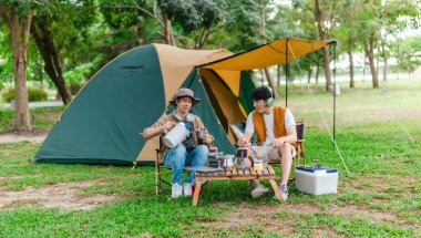 İki Asyalı erkek arkadaş doğayla çevrili kamp alanında dinlenmenin keyfini çıkarıyorlar. Etraflarında bir çadır kurulmuş ve teçhizat ile atmosfer rahatlığı, arkadaşlığı ve açık hava özgürlüğünü yansıtıyor.