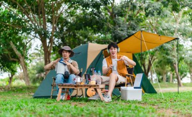 İki Asyalı erkek arkadaş doğayla çevrili kamp alanında dinlenmenin keyfini çıkarıyorlar. Etraflarında bir çadır kurulmuş ve teçhizat ile atmosfer rahatlığı, arkadaşlığı ve açık hava özgürlüğünü yansıtıyor.