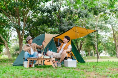 İki Asyalı erkek arkadaş doğayla çevrili kamp alanında dinlenmenin keyfini çıkarıyorlar. Etraflarında bir çadır kurulmuş ve teçhizat ile atmosfer rahatlığı, arkadaşlığı ve açık hava özgürlüğünü yansıtıyor.