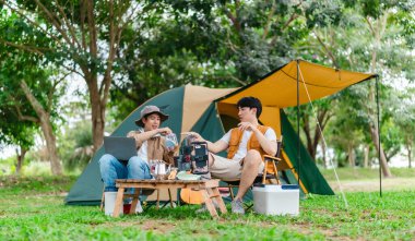 İki Asyalı erkek arkadaş doğayla çevrili kamp alanında dinlenmenin keyfini çıkarıyorlar. Etraflarında bir çadır kurulmuş ve teçhizat ile atmosfer rahatlığı, arkadaşlığı ve açık hava özgürlüğünü yansıtıyor.