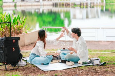 Asyalı bir çift göl kenarındaki piknik sırasında atıştırmalıkları paylaşırken keyifli bir anın tadını çıkarıyor. Yeşilliklerle çevrili sahne, doğadaki birlikteliğin rahatlatıcı ve neşeli atmosferini yakalıyor..