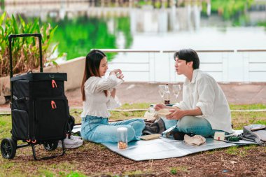 Asyalı bir çift göl kenarındaki piknik sırasında atıştırmalıkları paylaşırken keyifli bir anın tadını çıkarıyor. Yeşilliklerle çevrili sahne, doğadaki birlikteliğin rahatlatıcı ve neşeli atmosferini yakalıyor..