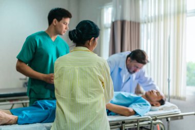 Endişeli bir kadın hastane yatağında yatan bir hastanın yanında tıbbi uzmanlarla konuşuyor. Olay yeri, klinik acil durum ortamında bir anlık iletişim ve destek görüntüsü yakalıyor..