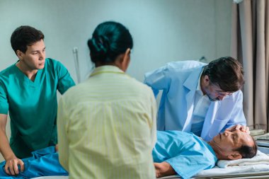 Endişeli bir kadın hastane yatağında yatan bir hastanın yanında tıbbi uzmanlarla konuşuyor. Olay yeri, klinik acil durum ortamında bir anlık iletişim ve destek görüntüsü yakalıyor..