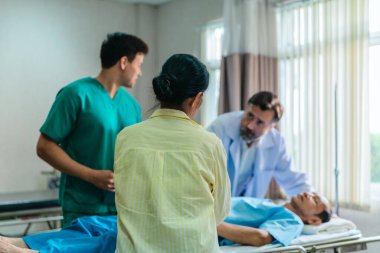 Endişeli bir kadın hastane yatağında yatan bir hastanın yanında tıbbi uzmanlarla konuşuyor. Olay yeri, klinik acil durum ortamında bir anlık iletişim ve destek görüntüsü yakalıyor..
