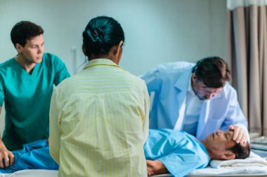 Endişeli bir kadın hastane yatağında yatan bir hastanın yanında tıbbi uzmanlarla konuşuyor. Olay yeri, klinik acil durum ortamında bir anlık iletişim ve destek görüntüsü yakalıyor..