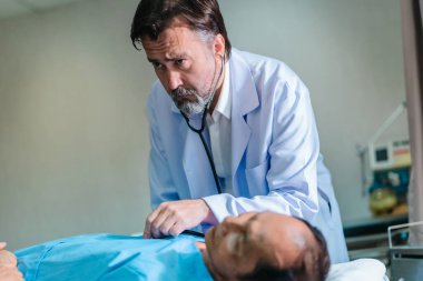 Başhekim hastanın hayati belirtilerini dikkatle incelerken hasta bir hastane yatağında uzanıyor ve klinik ortamda tıbbi bakım ve profesyonel sağlık hizmetlerini ön plana çıkarıyor..