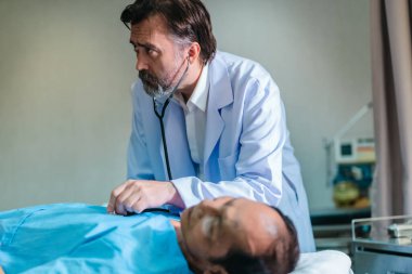 Başhekim hastanın hayati belirtilerini dikkatle incelerken hasta bir hastane yatağında uzanıyor ve klinik ortamda tıbbi bakım ve profesyonel sağlık hizmetlerini ön plana çıkarıyor..