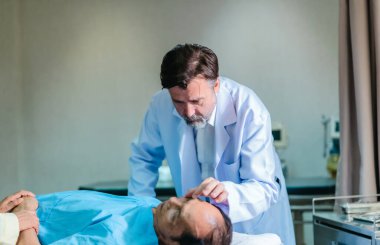 Başhekim hastanın hayati belirtilerini dikkatle incelerken hasta bir hastane yatağında uzanıyor ve klinik ortamda tıbbi bakım ve profesyonel sağlık hizmetlerini ön plana çıkarıyor..