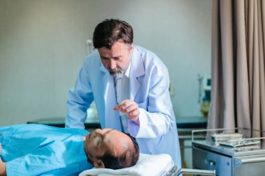 Başhekim hastanın hayati belirtilerini dikkatle incelerken hasta bir hastane yatağında uzanıyor ve klinik ortamda tıbbi bakım ve profesyonel sağlık hizmetlerini ön plana çıkarıyor..