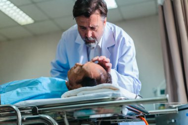 Başhekim hastanın hayati belirtilerini dikkatle incelerken hasta bir hastane yatağında uzanıyor ve klinik ortamda tıbbi bakım ve profesyonel sağlık hizmetlerini ön plana çıkarıyor..