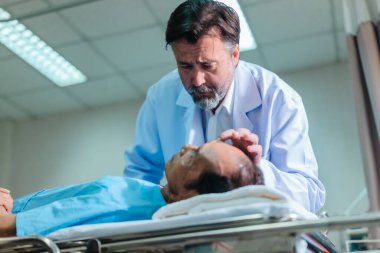 Başhekim hastanın hayati belirtilerini dikkatle incelerken hasta bir hastane yatağında uzanıyor ve klinik ortamda tıbbi bakım ve profesyonel sağlık hizmetlerini ön plana çıkarıyor..