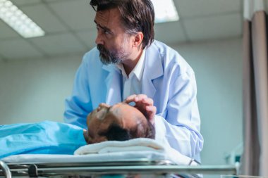 Başhekim hastanın hayati belirtilerini dikkatle incelerken hasta bir hastane yatağında uzanıyor ve klinik ortamda tıbbi bakım ve profesyonel sağlık hizmetlerini ön plana çıkarıyor..