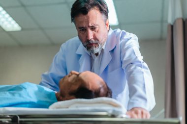 Başhekim hastanın hayati belirtilerini dikkatle incelerken hasta bir hastane yatağında uzanıyor ve klinik ortamda tıbbi bakım ve profesyonel sağlık hizmetlerini ön plana çıkarıyor..