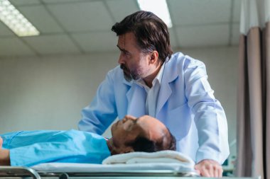 Başhekim hastanın hayati belirtilerini dikkatle incelerken hasta bir hastane yatağında uzanıyor ve klinik ortamda tıbbi bakım ve profesyonel sağlık hizmetlerini ön plana çıkarıyor..