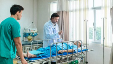 Kıdemli hekim hastane yatağında yatan hastayı muayene etmek için steteskop kullanırken, bir doktor dikkatlice gözlemliyor. Kurulum, profesyonel tıp ortamında acil durum ve takım çalışmasını iletiyor