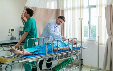 Kıdemli hekim hastane yatağında yatan hastayı muayene etmek için steteskop kullanırken, bir doktor dikkatlice gözlemliyor. Kurulum, profesyonel tıp ortamında acil durum ve takım çalışmasını iletiyor