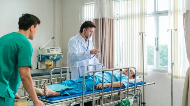 Kıdemli hekim hastane yatağında yatan hastayı muayene etmek için steteskop kullanırken, bir doktor dikkatlice gözlemliyor. Kurulum, profesyonel tıp ortamında acil durum ve takım çalışmasını iletiyor