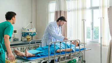Kıdemli hekim hastane yatağında yatan hastayı muayene etmek için steteskop kullanırken, bir doktor dikkatlice gözlemliyor. Kurulum, profesyonel tıp ortamında acil durum ve takım çalışmasını iletiyor