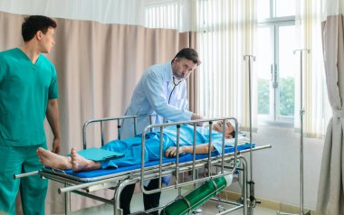 Kıdemli hekim hastane yatağında yatan hastayı muayene etmek için steteskop kullanırken, bir doktor dikkatlice gözlemliyor. Kurulum, profesyonel tıp ortamında acil durum ve takım çalışmasını iletiyor